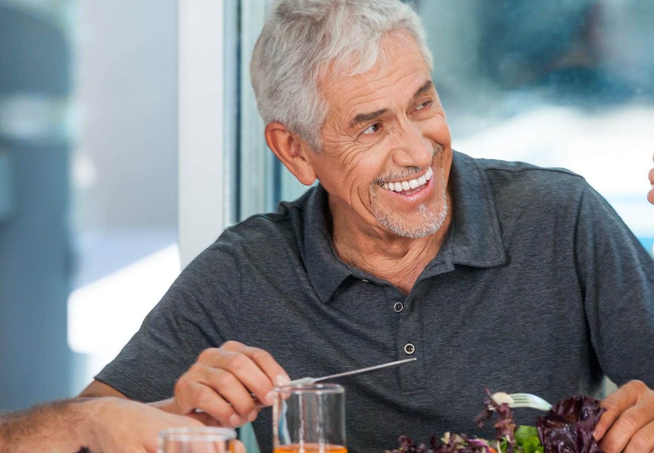 An older man with gray hair and a gray polo shirt is smiling while sitting at a table, holding a fork and eating a salad.