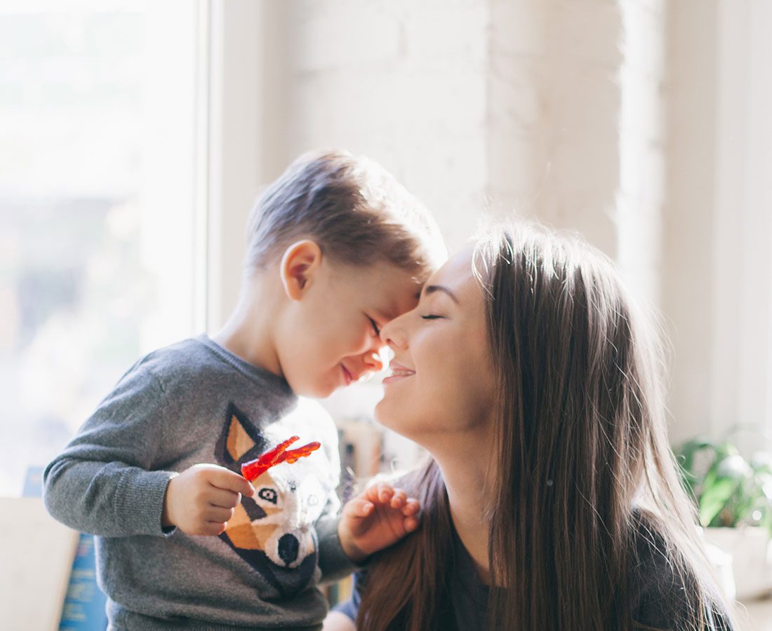 A young child and a woman smile with their faces touching gently in a sunlit room. The child wears a gray shirt with an animal graphic, and both appear joyful, sharing a tender and happy moment together.