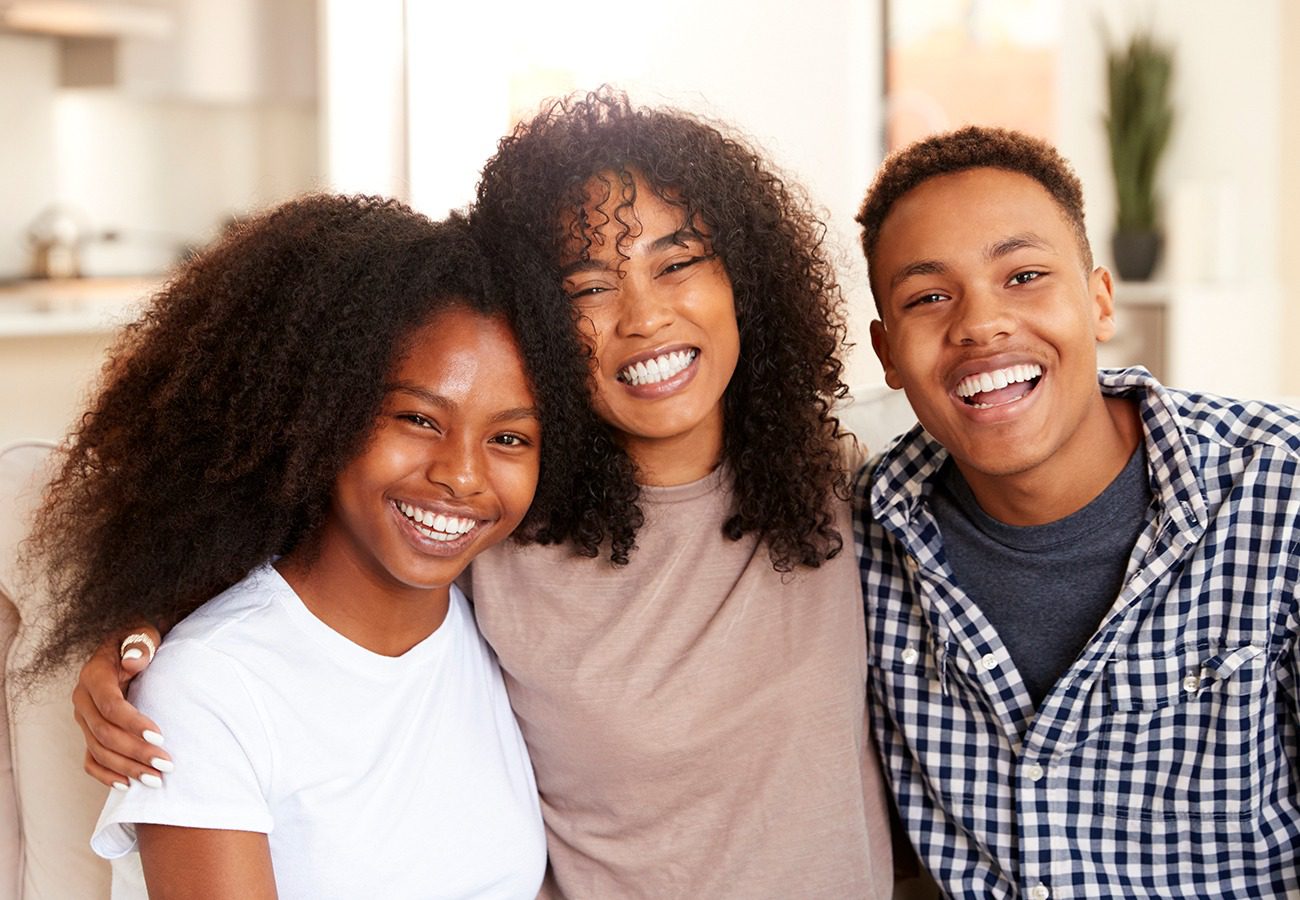 Three young adults sit close together indoors, smiling brightly at the camera. Two women and one man have curly hair and casual clothes, and they appear happy and relaxed in a home setting.