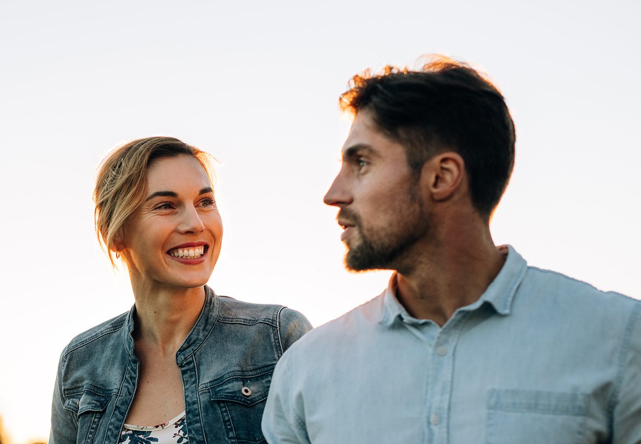 A woman smiling at a man outdoors at sunset, both wearing casual denim shirts, with a clear sky in the background.