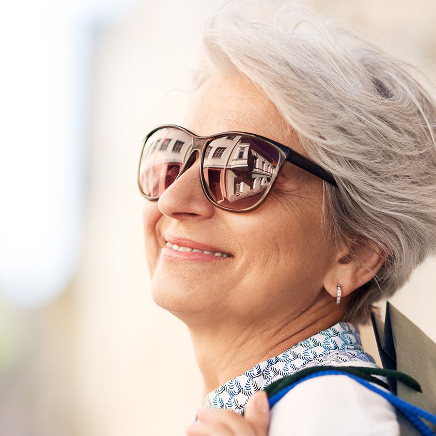 An older woman with short gray hair smiles while wearing large sunglasses and earrings. Her sunglasses reflect buildings, and she appears to be outdoors on a sunny day, carrying a shopping bag over her shoulder.