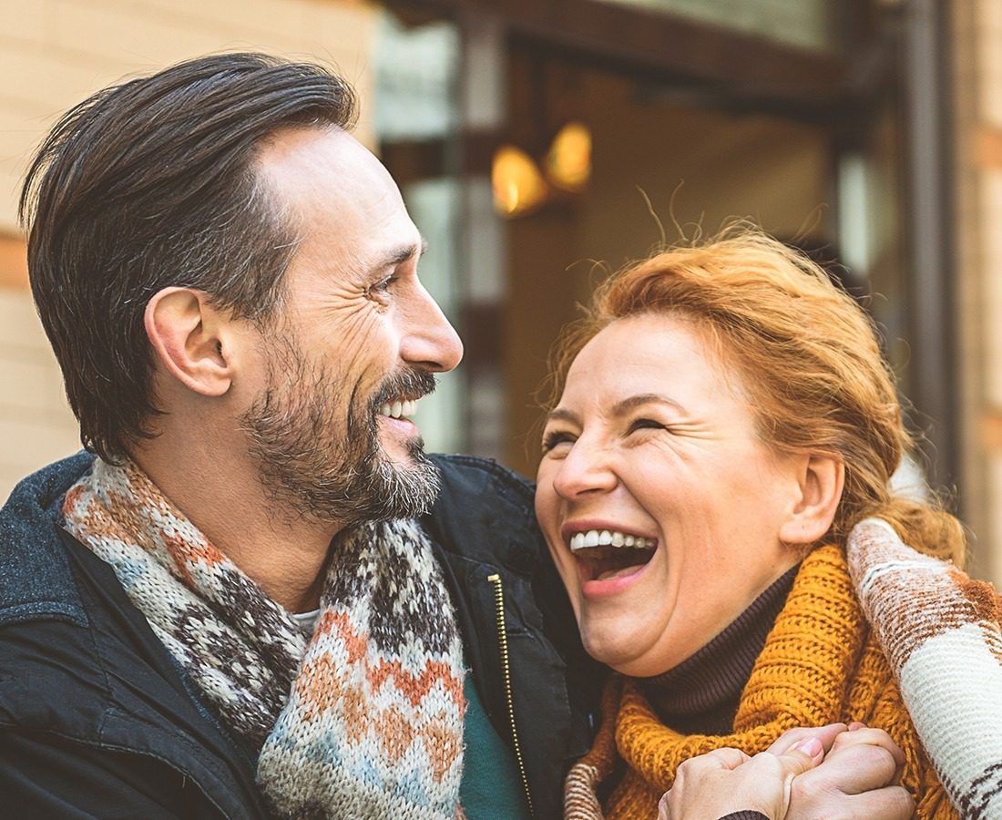 A smiling man and woman wearing winter scarves and coats stand close together outdoors, laughing and looking at each other warmly.