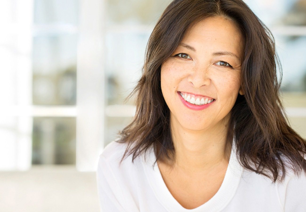 A woman with medium-length dark hair and a white shirt smiles warmly at the camera, with a softly blurred background of windows and natural light.