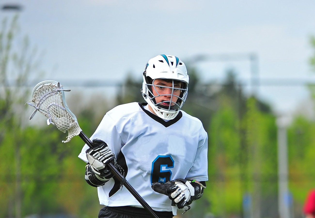 A lacrosse player wearing a white jersey with the number 6, white helmet, and gloves runs on a field holding a lacrosse stick, with green trees and a blurred background behind him.