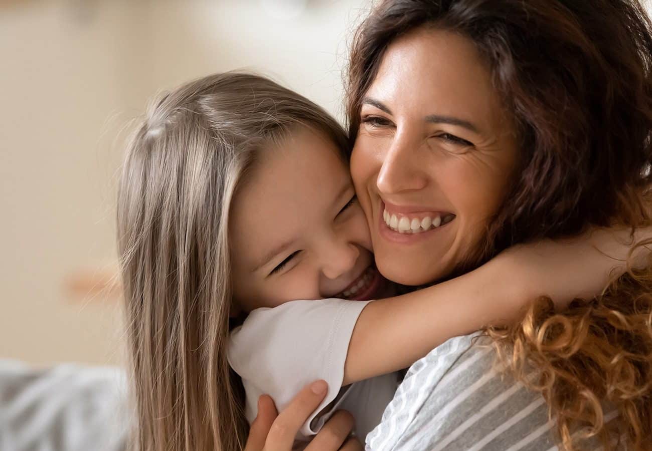 A young girl with long brown hair hugs a smiling woman, possibly her mother. Both look happy and are laughing, with their faces close together in an affectionate embrace.