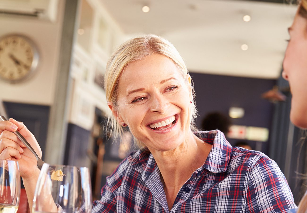 A smiling woman with blonde hair, wearing a plaid shirt, sits at a table in a restaurant, holding a fork and talking to someone off-camera. A wine glass and a clock on the wall are visible in the background.