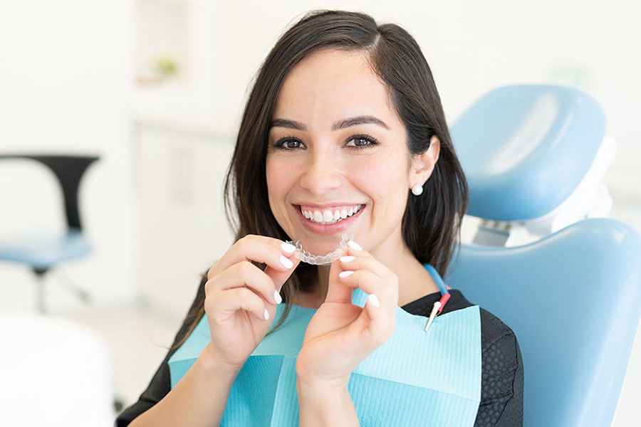 A woman sitting in a dental chair smiles while holding a clear orthodontic aligner. She wears a dental bib and appears to be at a dental clinic.