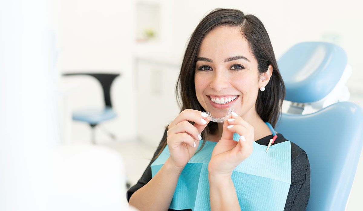 A woman sitting in a dental chair smiles while holding a clear orthodontic aligner. She wears a dental bib and appears to be at a dental clinic.