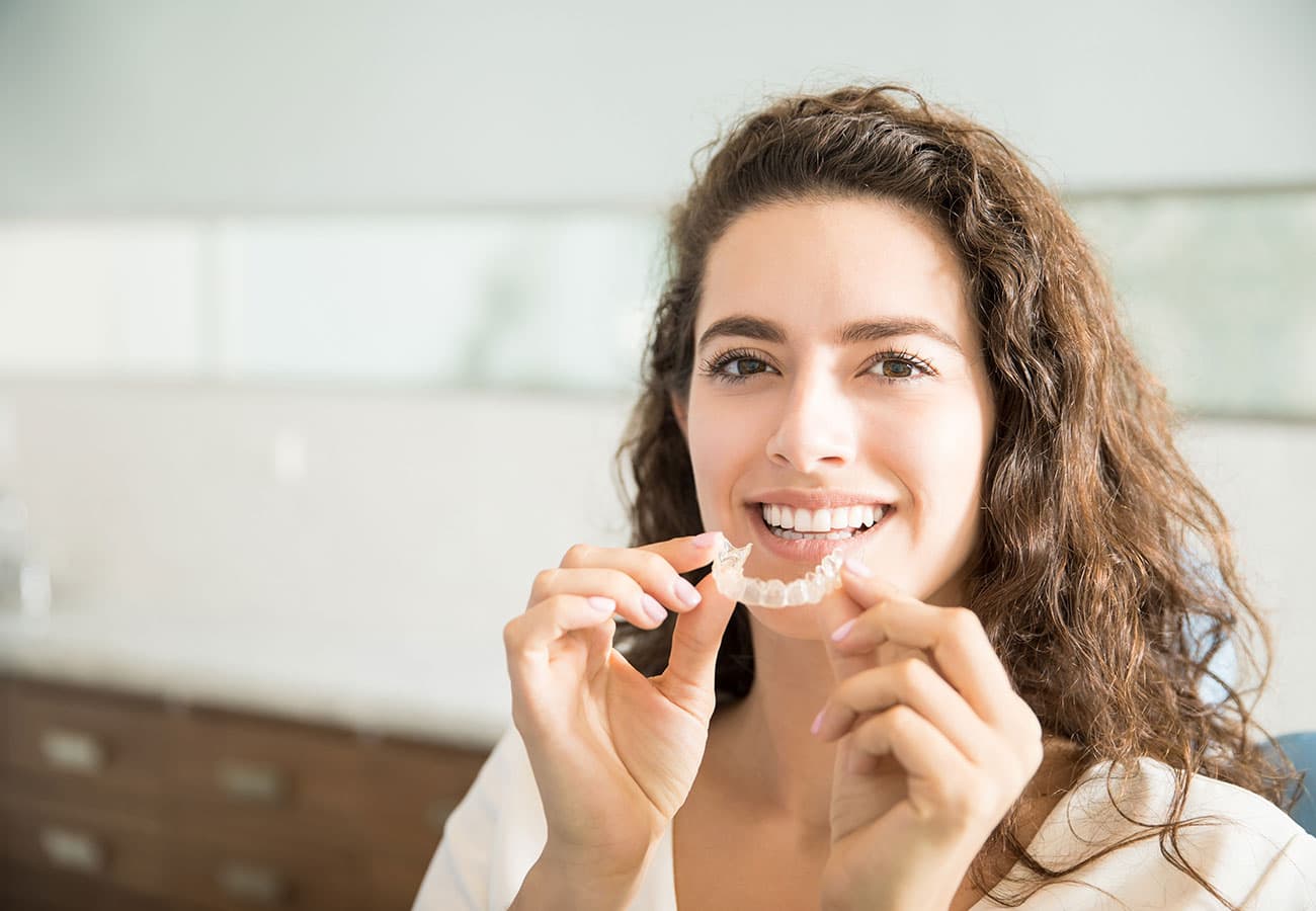 A woman with long curly hair smiles while holding a clear dental aligner near her mouth, ready to place it on her teeth. She is indoors in a bright, modern room.